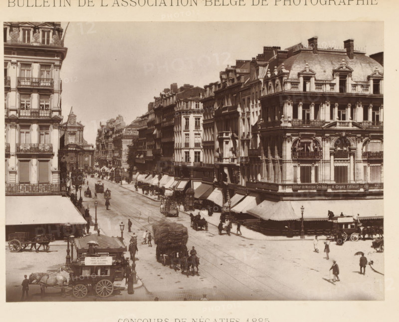 Street view in Brussels, page, photomechanical print, inscription: 'Concours de négatifs 1885', photographer: Henri Tournai, Brussels, cliché maker: workshop of Adolphe Braun & Cie., Dornach, c. 1880 - in or before 1885, printer (person), Mulhouse, paper, Woodburytype, height 118 mm x width 157 mm, Print on unnumbered page., street, A wide street scene shows multiple ornate multi story buildings with decorative façades and balconies lining both sides, a corner building with a rounded roof and sculpted window surrounds visible on the right, rows of awnings extending over shopfronts beneath the upper floors, and a distant churchlike building with a pediment visible along the left side. The street is populated with horse-drawn carriages and wagons, drivers and pedestrians walking or standing, a large load of bundled material on a cart moving down the road, several street lamps along the pavement, and small kiosk or booth near the foreground curb. The image is monochrome with a slightly fa...