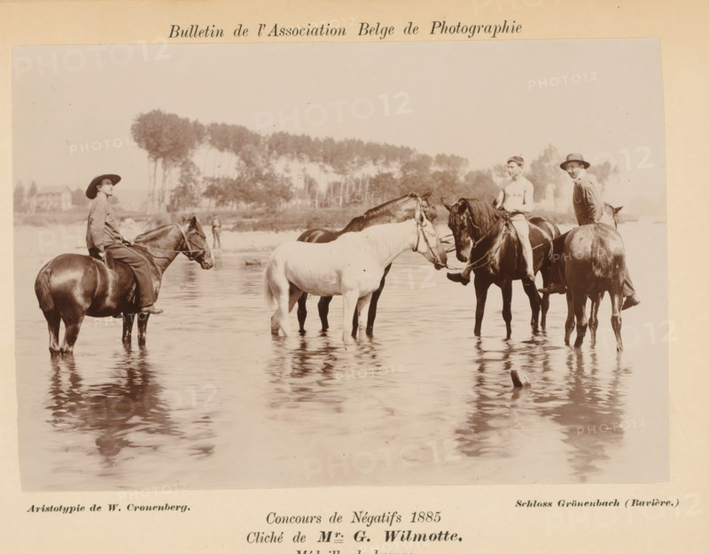 Riders in a river, photograph, page, inscription: 'Concours de Négatifs 1885, Médaille de bronze.', photographer: Georges Wilmotte, printer (person): Wilhelm Cronenberg, Grönenbach, c. 1880 - in or before 1885, printer (person), Bavaria, paper, pasteboard, baryta paper, daylight collodion silver print, height 104 mm x width 154 mm, Photo on unnumbered page., horse, riding a horse, ass, or mule, rider, horseman, river, A sepia photograph shows several people on horseback standing in shallow water with reflections on the surface; one person on the left wears a wide-brimmed hat and loose clothing while sitting on a dark horse, a shirtless rider sits on a dark horse near the center, another person in a hat and jacket sits on a horse to the right, and a light-colored horse and a dark horse stand together between them; trees and distant buildings form a blurred background across the horizon above the group.