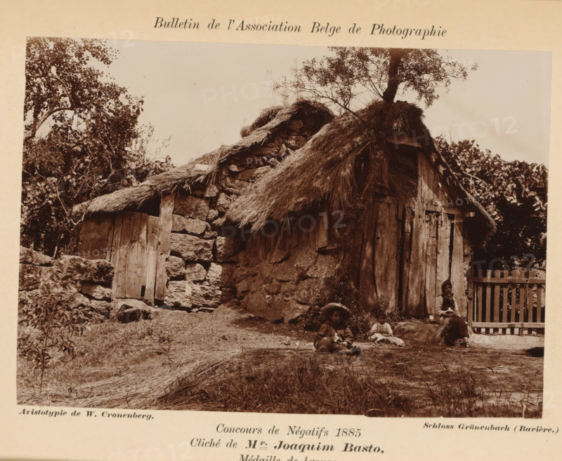 House in Vizela, Portugal, Une cabane à Vizeila, photograph, page, inscription: 'Concours de Négatifs 1885, Médaille de bronze.', photographer: Joaquim Basto, Vizela, printer (person): Wilhelm Cronenberg, Grönenbach, c. 1880 - in or before 1885, printer (person), Bavaria, Braga, paper, pasteboard, baryta paper, daylight collodion silver print, height 102 mm x width 147 mm, Photo on unnumbered page., hut, cabin, lodge, A sepia photograph showing a rustic stone and wood cottage with a thatched roof, surrounded by trees and foliage, a wooden door set into a stone wall, a wooden fence to the right, and three people seated on the ground and a low stone ledge near the cottage, one wearing a wide-brimmed hat and another holding a bundle of straw, with scattered straw and grassy ground in the foreground.