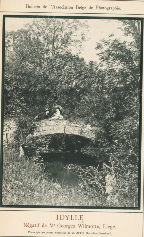Man and woman on a bridge over the Berwinne, Idylle, page, photomechanical print, photographer: Georges Wilmotte, Liège, cliché maker: Wilhelm Otto, Brussels, c. 1881 - in or before 1886, printer (person), paper, collotype, photoprint, height 160 mm x width 116 mm, Print on unnumbered page., brook, bridge, lovers' meeting, Berwinne, A monochrome photograph framed by a printed page shows a stone arch bridge over a dark, narrow stream surrounded by dense foliage and tall trees; two people sit atop the bridge near its center, one holding an open parasol while the other leans toward them, and grasses and shrubs grow along the banks and foreground.