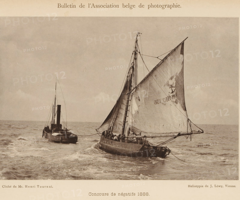Seascape with sailing ship and tugboat, page, photomechanical print, inscription: 'Concours de négatifs 1888., Médaille de Vermeil.', photographer: Henri Tournai, cliché maker: Josef Löwy, Vienna, c. 1883 - in or before 1888, printer (person), Lower Austria, paper, pasteboard, heliotype, height 115 mm x width 168 mm, Print on unnumbered page., sailing-ship, sailing-boat, sea (seascape), tug, pusher tug, A sepia-toned photograph showing two boats on a choppy sea, one closer with a large triangular sail raised and visible rigging, the hull rounded and crew members aboard, and another boat behind with a vertical smokestack and mast; the sky above is mostly clear with subtle cloud texture and the water shows waves and wake around the vessels.