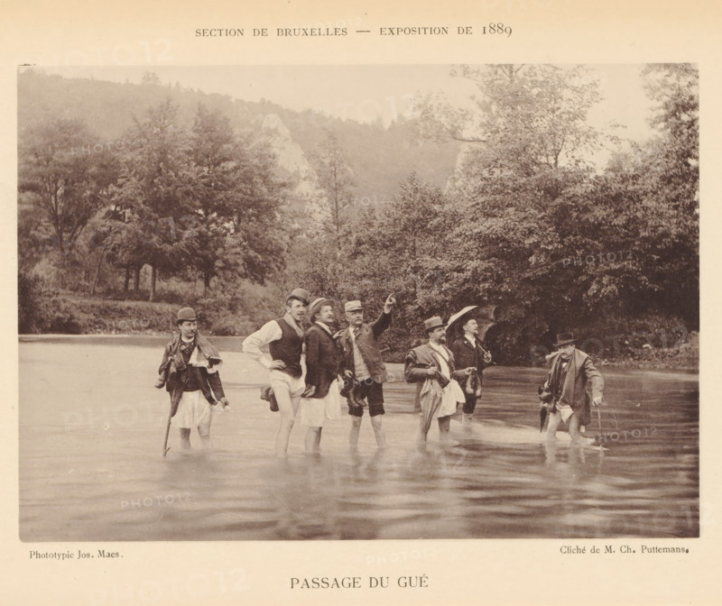 Passage du Gué, Group of men in the river Lesse, page, photomechanical print, inscription: 'Section de Bruxelles - Exposition de 1889', photographer: Charles Puttemans, Lesse, cliché maker: Joseph Maes, Antwerp, c. 1883 - in or before 1888, printer (person), Maas, paper, collotype, photoprint, height 115 mm x width 164 mm, Print on unnumbered page., adult man, crossing a river, A sepia photograph shows a group of men standing waist-deep in a body of water near a wooded shoreline, wearing period clothing including jackets, hats, and walking sticks; some men hold umbrellas or bags, one man points outward while others look in various directions, and trees and a hillside form the background.