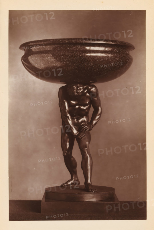 Porphyry bowl supported by a bronze statue of Hercules, from the collection of Christopher Beckett Denison, page, photomechanical print, number: '807', after sculpture by anonymous, c. 1880 - in or before 1885, after sculpture by, paper, Woodburytype, height 186 mm x width 122 mm, Print on seventh unnumbered page after page 78., (story of) Hercules (Heracles), dish, plate, saucer, piece of sculpture (precious metals, arts), A sepia photograph of a sculpture showing a muscular male figure standing on a round stepped base, the figure slightly bent forward with head lowered and one arm across the groin while the other grasps the wrist, supporting a large shallow bowl balanced on the back of the neck and shoulders; the bowl has a speckled surface and a rounded rim, the figure is detailed with defined musculature and textured hair, and the background is plain with a light-to-dark gradient.