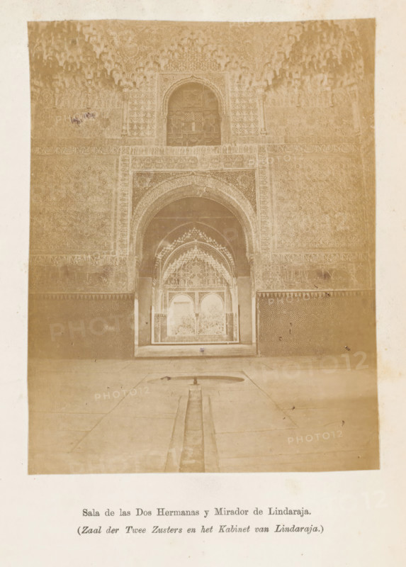 Sala de las Dos Harmanas y Mirador de Lindaraja, Zaal der Twee Zusters en het Kabinet van Lindaraja, Patio van Lindaraja gezien door de Zaal der Twee Zusters in het Alhambra, photograph, page, Granada, c. 1875 - in or before 1880, photographer, Andalusia, paper, albumen print, height 128 mm x width 99 mm, interior, representation of a building, Alhambra, A sepia-toned photograph of an ornate interior showing a large arched doorway with layered horseshoe and scalloped arches leading into a smaller archway and a recessed alcove, all richly decorated with geometric and floral carved plasterwork and repeating patterns; a narrow channel runs along a tiled floor toward the doorway, ending in a shallow basin, and the walls flanking the arches display dense patterned panels and decorative friezes, with a vaulted muqarnas-like ceiling above the central arch.