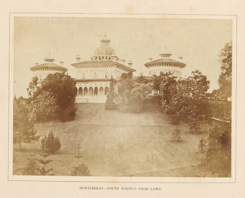 View of a portico of the Palácio de Monserrate near Sintra, Montserrat - South portico from lawn, photograph, page, Sintra, c. 1865 - in or before 1870, photographer, paper, albumen print, height 143 mm x width 197 mm, palace, gate, entrance, Palácio de Monserrate, A sepia-toned photograph showing a large ornate building with a central dome and two round towers on either side, all topped with decorative roofs and small cupolas, a portico with a row of arched openings across the front, balustrades beneath the arches, and carved architectural details along the cornices; the building is set behind a gently sloping grassy lawn bordered by dense trees and shrubs of varying heights that partially obscure the lower façade, with scattered small plantings visible on the lawn and a pale sky above.