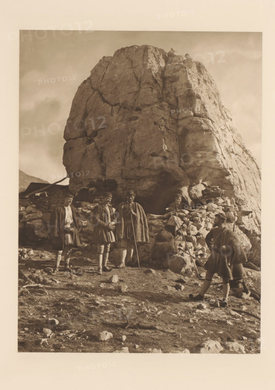 Les bergers de Parnasse et leurs hôtes, Shepherds on Parnassus, page, photomechanical print, photographer: Frédéric Boissonnas, Parnassus, cliché maker: Meisenbach, Riffarth & Cie., Berlin, c. 1905 - in or before 1910, paper, photoaquatint, height 290 mm x width 224 mm, Photo on page 96., herding, herdsman, herdswoman, shepherd, shepherdess, cowherd, A large rocky outcrop fills the center and upper portion of the image, with rough, cracked surfaces and light and shadow defining its contours. At the base of the rock is a low stone wall or pile of stones forming a rough shelter area. Five people wearing heavy cloaks and traditional-looking garments stand and sit near the stone wall; some hold long staffs or walking sticks and one appears seated among the stones. The ground in the foreground is uneven and strewn with rocks and small boulders. The overall image has a monochrome, sepia-toned appearance.