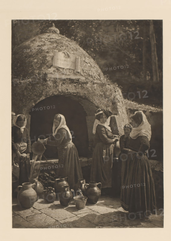 Women at a well in Gastouri on Corfu, Le puits de Gastouri à Corfou, page, photomechanical print, photographer: Frédéric Boissonnas, Gastouri, cliché maker: Meisenbach, Riffarth & Cie., Berlin, c. 1905 - in or before 1910, paper, photoaquatint, height 288 mm x width 208 mm, Photo on page 12., adult woman, well, A monochrome photograph showing a rounded stone structure with an arched opening and a carved plaque mounted near its top, a group of women wearing long dresses and head coverings gathered in front of the opening, one woman tipping a pitcher to pour water into a basin while several earthenware jugs and pots of various shapes sit on the tiled ground in front of them, a wooded background visible beyond the stone structure.