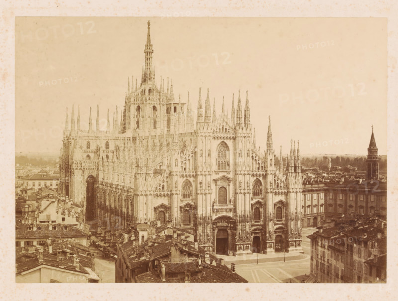 View of the Milan Cathedral, photograph, signature: 'Fotografia Artistica P. Pozzi Milano', photographer: Pompeo Pozzi, publisher: William Spooner, c. 1860 - c. 1865, pasteboard, paper, albumen print, height 256 mm x width 358 mm, Lombardy, Milan, Cathedral (Milan), A wide sepia-toned view of an ornate Gothic cathedral facade dominated by tall spires, pointed arches, intricate tracery, and carved statues, with detailed doorways and large stained glass window openings; surrounding the cathedral are densely packed old buildings with tiled roofs and narrow streets, and a square in front of the cathedral with a paved surface; in the distance a tall tower with a conical roof rises above lower rooftops, and the sky above is pale and mostly clear.