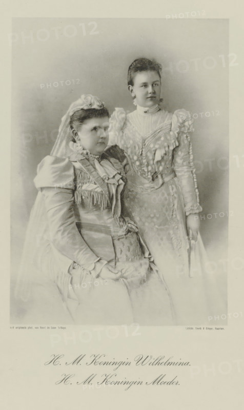 Portrait of Emma, Queen Regent of the Netherlands, and Wilhelmina, Queen of the Netherlands, photomechanical print, Portrait of Emma and Wilhelmina. Their names and titles in the lower margin., photographer: Henri de Louw, cliché maker: Emrik & Binger, Haarlem, 1897, paper, collotype, photogravure, height 264 mm x width 195 mm, The title mentioned here refers to the highest title obtained by the sitter, not necessarily to the title the sitter bears in this image., Person, Emma (Queen consort of the Netherlands), Wilhelmina of the Netherlands, A sepia-toned studio portrait showing two women in ornate dresses; one woman is seated wearing a high-necked gown with layered ruffles, a veil, and a headpiece of flowers, her hands resting in her lap, while the other woman stands slightly behind and to the side wearing a high-collared gown with puffed sleeves, lace and embroidered detailing, and she holds a folded fan at her side; both have hair styled up and neutral facial expressions, and the b...