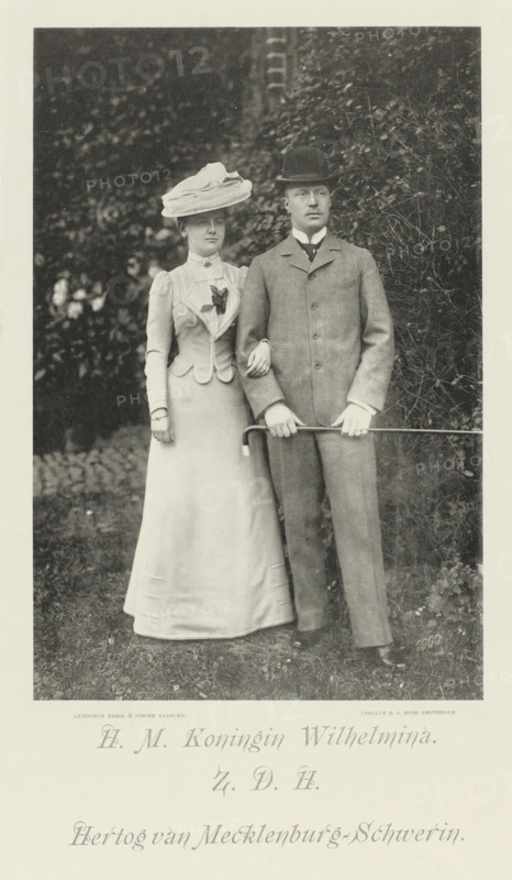 Portrait of Wilhelmina, Queen of the Netherlands, and Hendrik of Mecklenburg-Schwerin, photomechanical print, Portrait of Wilhelmina and Hendrik. Their names and titles in the lower margin., cliché maker: Emrik & Binger, Haarlem, publisher: Nicolaas Jan Boon, Amsterdam, 1900, photographer, paper, collotype, photogravure, height 346 mm x width 229 mm, The title mentioned here refers to the highest title obtained by the sitter, not necessarily to the title the sitter bears in this image., Person, Hendrik (Prince of the Netherlands (1876-1934)), Wilhelmina of the Netherlands, A black-and-white photograph of a woman and a man standing outdoors against a background of dense foliage, the woman on the left wearing a long light-colored dress with a fitted jacket, a large wide-brimmed hat with layered fabric, a decorative floral detail on her chest, and her arm linked through the man's arm; the man on the right wearing a three-piece suit with a jacket buttoned at the front, a collared shirt wit...