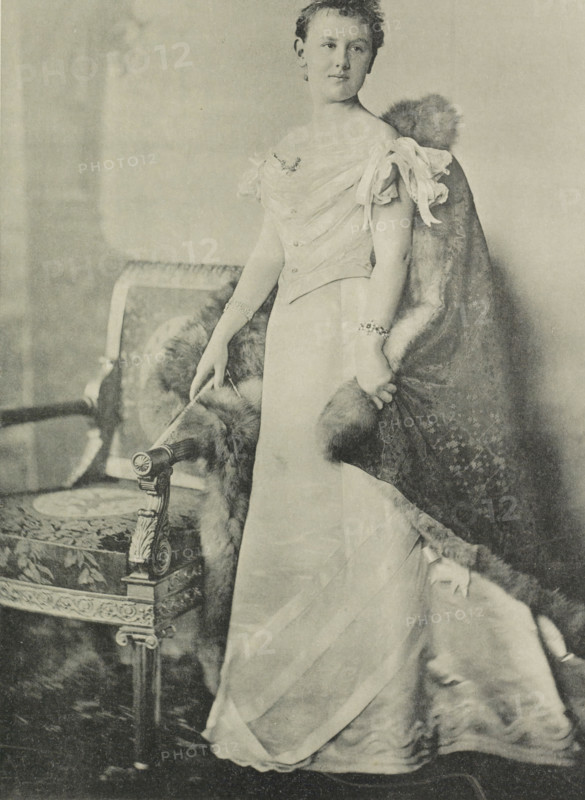 Portrait of Wilhelmina, Queen of the Netherlands, photomechanical print, Portrait of Wilhelmina. She stands next to a chair. In the lower margin her name and title., photographer: Heinrich Wilhelm Wollrabe, clichémaker: anonymous, Netherlands, 1897, paper, collotype, photoprint, height 347 mm x width 217 mm, The title mentioned here refers to the highest title obtained by the sitter, not necessarily to the title the sitter bears in this image., Person, Wilhelmina of the Netherlands, A sepia-toned portrait of a woman standing beside an ornately carved upholstered chair, wearing a floor-length gown with an off-the-shoulder bodice and delicate draped fabric at the sleeves, a fur-trimmed cloak draped over one shoulder and trailing toward the floor, bracelets on both wrists, one hand resting on the chair arm and the other holding the fur, her hair styled up, and the chair featuring decorative carved details and patterned upholstery.