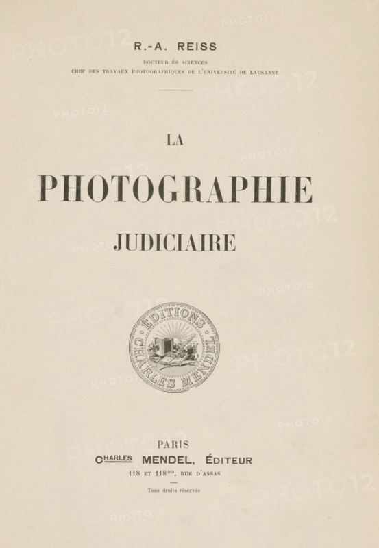 La photographie judiciaire, book, name: 'Reinders', writer (clerical worker): Rodolphe Archibald Reiss, publisher: Charles Mendel, Paris, 1903, writer (clerical worker), textile materials, pasteboard, paper, linen (material), printing, marbled paper, gelatin silver print, autotype, developing gelatin silver print, height 248 mm x width 185 mm x thickness 25 mm, A vertical cream-colored page with centered dark serif headings and a circular emblem lower on the page; the top shows a small bold heading, beneath it narrower lines of smaller text, then a thin horizontal rule, followed by a single short word in medium-sized serif; below that a large bold word in wide spaced capital letters followed by another medium bold word; lower on the page is a round decorative seal with an inner illustration and concentric patterned border; near the bottom are additional lines of smaller serif text and a small centered short line above the very bottom of the page.