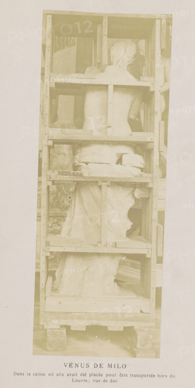 Venus de Milo in a transport box, seen from the back, Vénus de Milo, photograph, page, inscription: 'Dans la caisse, vue de dos', c. 1861 - in or before 1871, photographer, paper, albumen print, height 179 mm x width 67 mm, sculpture, (story of) Venus (Aphrodite), A tall wooden crate with horizontal and vertical planks holds a fragmented marble statue shown from the back, with a torso and draped lower body visible, the statue’s head turned slightly to one side and supported within the crate, and wooden blocks and padding placed around the sculpture; the crate rests on a pallet and the photograph has a light, faded tone.