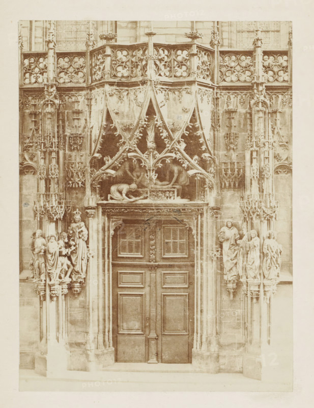 Entrance portal of the Saint Lawrence Chapel of the Strasbourg Cathedral, photograph, page, photographer: Charles David Winter, Strasbourg (France), c. 1851 - in or before 1861, paper, albumen print, height 148 mm x width 110 mm, sculpture (relief, sculpture), parts of church exterior and annexes: portal, Notre-Dame (Strasbourg), A tall ornate doorway set into a stone façade with carved panels and a central wooden double door framed by decorative molding, above which sits an elaborate canopy of pointed arches and foliated tracery with sculpted figures and relief work; flanking the entrance are clustered vertical columns with pinnacles and niches containing statues of robed figures standing on small plinths, and additional carved architectural ornamentation fills the upper facade with repeating quatrefoil patterns and intricate stone latticework.