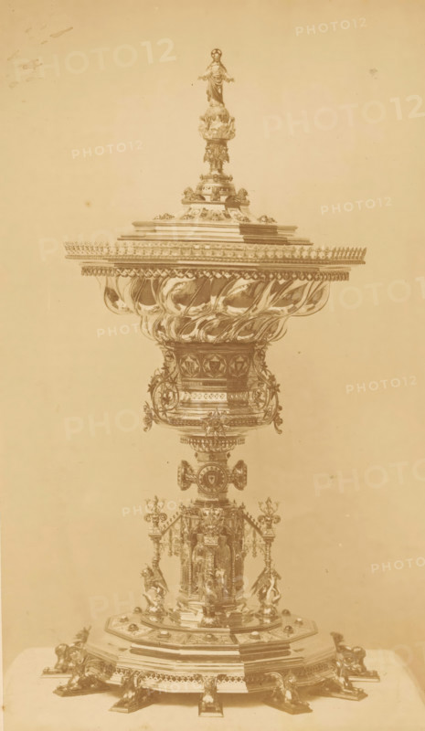 Ciborie, photograph, page, c. 1858 - in or before 1868, photographer, paper, albumen print, height 218 mm x width 126 mm, ciborium, A tall ornate metal vessel with a wide flared bowl and a domed lid topped by a small standing figure, featuring elaborate scrolling and floral ornamentation along its rounded body and pedestal, supported by a detailed base with sculpted figures and decorative feet, all shown in a monochrome sepia tone against a plain background.