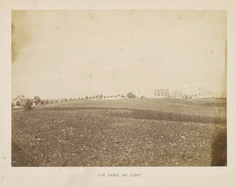 View of the observatory in Besançon, Vue prize de l'est, photograph, page, photographer: M. Lebeuf, Besançon, c. 1882 - in or before 1892, M. Lebeuf, photographer, paper, albumen print, height 160 mm x width 221 mm, observatory (, astronomy), meteorological observatory, Observatoire de Besançon, A wide sepia photograph showing a plowed field in the foreground with visible soil clumps and scattered stones, a gently sloping strip of grass and sparse vegetation above the field, and a low horizon line dotted with trees and small buildings including several houses and larger classical-style structures with columns and pitched roofs; the sky occupies much of the upper area and appears pale and empty with a few faint spots and blemishes on the photographic surface.