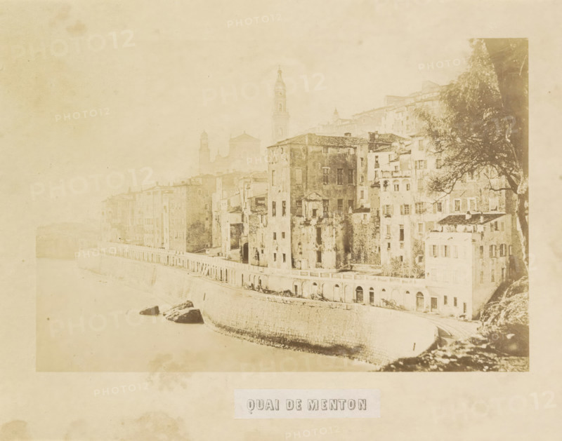 Quai de Menton, View of the quay of Menton, photograph, page, photographer: Louis-Alphonse Davanne, Menton, c. 1852 - in or before 1862, Provence-Alpes-Côte d'Azur, paper, albumen print, height 122 mm x width 182 mm, façade (of house or building), quay, A faded sepia photograph shows a riverside view with a stone embankment curving along the water and tall, weathered buildings rising directly from the embankment, their facades marked by windows, balconies, and patches of wear; a tower and church silhouettes are visible behind the row of buildings, and a large tree with spreading branches appears on the right side near the structures, while rocks rest near the water's edge and the overall image has an aged, grainy texture.