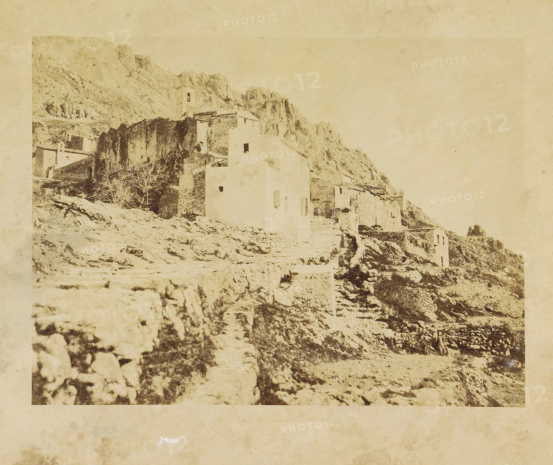 Ste. Agnes, View of Sainte-Agnès, photograph, page, photographer: Louis-Alphonse Davanne, Sainte-Agnès, c. 1852 - in or before 1862, Provence-Alpes-Côte d'Azur, paper, albumen print, height 137 mm x width 198 mm, Photo for page 40., city-view in general, 'veduta', Sainte-Agnes, A faded, sepia-toned photograph of a rocky hillside with a cluster of stone buildings and ruins built into the slope, showing rough stone walls, stairways, and terraced paths leading upward; one taller building with a small squared tower and simple rectangular openings is visible among lower structures, and sparse leafless trees and scattered rocks are present on the uneven terrain beneath a pale, textured sky.