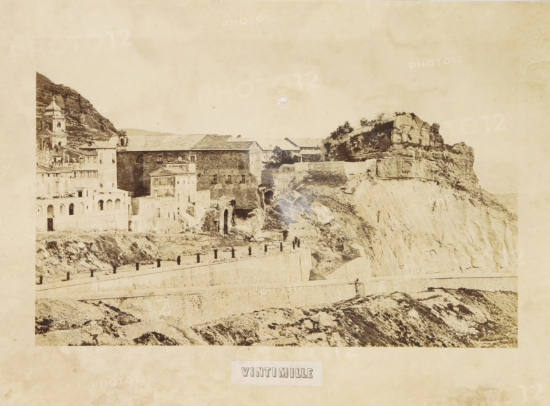 View of Ventimiglia, Vintimille, photograph, page, photographer: Louis-Alphonse Davanne, Italy, c. 1852 - in or before 1862, paper, albumen print, height 125 mm x width 197 mm, city-view in general, 'veduta', Ventimiglia, A sepia-toned photograph shows a cliffside town with stone buildings perched along steep rocky terrain, including a multi-story structure with arched openings and a taller tower with a domed roof on the left, a long stone wall running horizontally across the lower part of the scene with evenly spaced short posts along its top, rough rocky slopes and terraces beneath the wall, and large layered rock formations and vegetation on the right where the cliff face drops away.