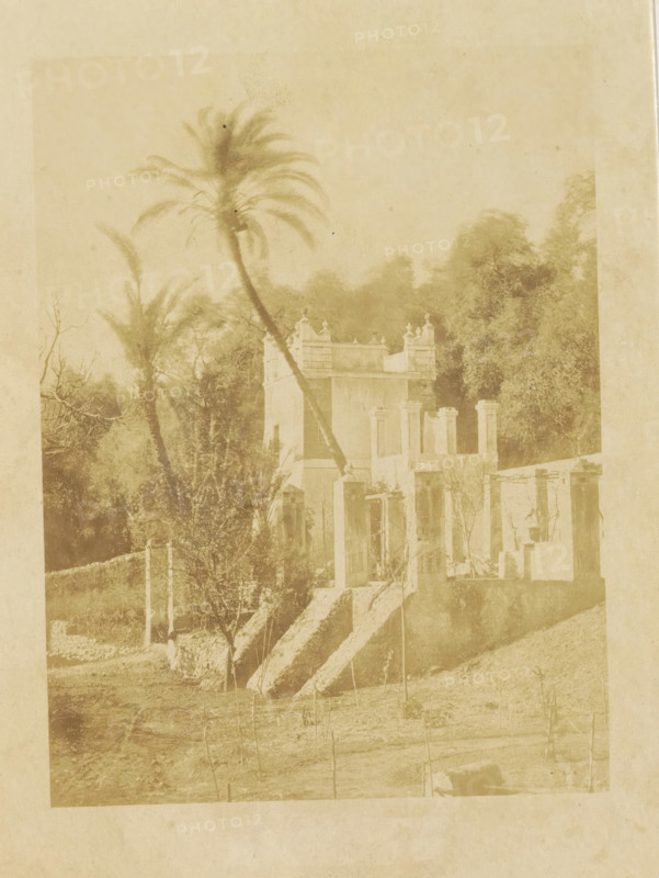 Jardins Moreno Bordighera, Part of the Moreno Gardens in Bordighera, photograph, page, photographer: Louis-Alphonse Davanne, Bordighera, c. 1852 - in or before 1862, paper, albumen print, height 176 mm x width 132 mm, Photo for page 51., garden, Ventimiglia, A faded sepia photograph showing a small building with ornate parapets and pillars, a set of worn stone steps leading up to doorways, and partial ruined walls; a tall, leaning palm tree rises beside the building with fronds splaying at the top, and additional trees and dense foliage fill the background; the foreground shows a rough grassy area with sparse young stakes or saplings and a low stone wall segment, all rendered in soft, aged tones.