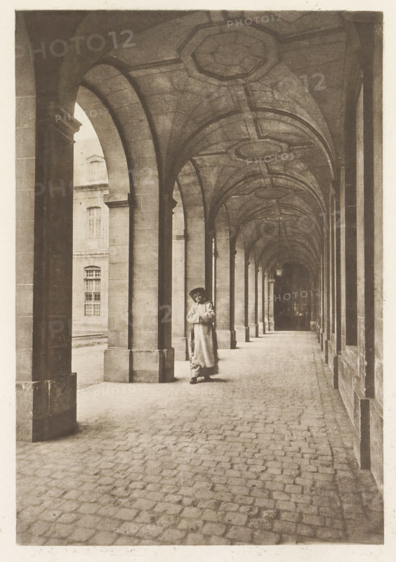 Monk in an arcade, page, photomechanical print, photographer: Henri Magron, France, cliché maker: Paul Dujardin, c. 1885 - in or before 1896, paper, photoaquatint, height 206 mm x width 140 mm, monk(s), friar(s), arcade, architecture, A long covered stone arcade with a vaulted ceiling and repeating arches receding into the distance, stone columns on one side and a wall with window openings on the other, a cobblestone floor, and a person standing near one of the central columns wearing a long coat and a hat while holding something in front of their chest.