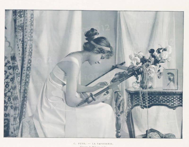 Woman behind a loom, La tapisserie, page, photomechanical print, name: 'Jean Malvaux PH', photographer: Constant Puyo, cliché maker: Malocuy et Cie, cliché maker: Malvaux et Cie, c. 1889 - in or before 1899, paper, autotypie, height 150 mm x width 210 mm, weaving-loom, A seated woman in a light sleeveless dress leans forward, her hair wrapped with a ribbon and gathered in a loose bun, as she works with a long handled tool over a frame; beside her an ornate table holds a vase of mixed flowers and a framed portrait, and yarn and cords hang and lie near the table; patterned drapery and plain curtains form the background.