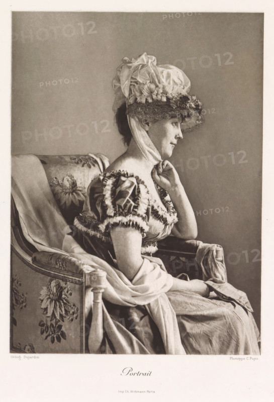 Portrait, Portrait of an unknown woman with a hat in a chair, page, photomechanical print, photographer: Constant Puyo, France, cliché maker: Paul Dujardin, printer (person): Charles Wittmann, Paris, c. 1886 - in or before 1896, printer (person), paper, photoaquatint, height 211 mm x width 153 mm, adult woman, head-gear: hat, chair, A sepia-toned portrait of a seated woman in profile facing right, wearing a large ornate bonnet with ruffled fabric and lace, a ribbon tied beneath her chin, and a dress with puffed sleeves trimmed with ruffles; she rests one hand lightly against her neck and her other hand lies in her lap, while she sits on a patterned upholstered chair draped with fabric and a long flowing skirt cascades over the seat and armrest.