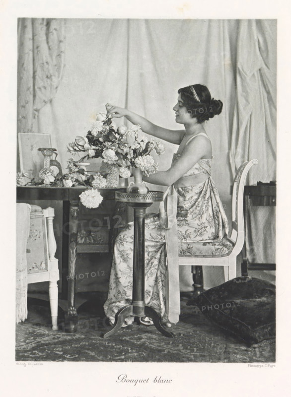 Flower arranging woman in an interior, Bouquet blanc, page, photomechanical print, photographer: Constant Puyo, France, cliché maker: Paul Dujardin, printer (person): Charles Wittmann, Paris, c. 1886 - in or before 1896, printer (person), paper, photoaquatint, height 218 mm x width 165 mm, adult woman, arranging flowers, interior, representation of a building, A seated woman in a patterned dress with a ribbon tied around her waist arranges a large bouquet of flowers on a wooden table, her hair pinned up with a headband and her profile visible as she reaches toward the blooms; the table holds various vases and scattered blossoms, and an ornate chair with a cushioned seat supports her while a small round pedestal table stands in front; draped fabric hangs in the background and a patterned rug covers the floor, with a dark cushion or piece of fabric resting nearby.