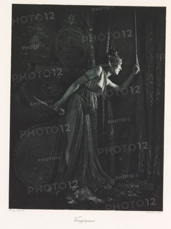 Woman with a knife in hand, depicting the Revenge, Vengeance, page, photomechanical print, photographer: Constant Puyo, France, cliché maker: Paul Dujardin, printer (person): Charles Wittmann, Paris, c. 1886 - in or before 1896, printer (person), paper, photoaquatint, height 216 mm x width 164 mm, adult woman, curtains, tools, aids, implements, crafts and industries: knife, A monochrome image shows a woman in a flowing dress leaning forward and parting a heavy patterned curtain with one hand while holding a curved dagger behind her back with the other; she wears an ornate headpiece with round decorative elements and beaded straps across her shoulders and bodice, and a round decorated shield rests near her side while a feathered object lies on the floor at her feet.