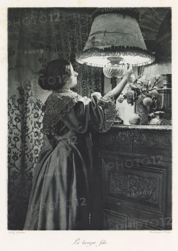 Woman turns on a lamp, La lampe file, page, photomechanical print, photographer: Constant Puyo, France, cliché maker: Paul Dujardin, printer (person): Charles Wittmann, Paris, c. 1886 - in or before 1896, printer (person), paper, photoaquatint, height 218 mm x width 160 mm, adult woman, lighting, lamps, A woman in a long dark dress with a lace collar and sleeves stands in profile reaching up to adjust a large fringed lampshade; her hair is pulled back in a bun and she faces the illuminated interior of the lamp. The lamp has a decorative base and sits on a carved wooden surface beside a vase of flowers and other small objects. Behind her is a patterned textile or wallpaper with floral motifs. The scene is rendered in grayscale with soft lighting highlighting the woman's face and the lamp area.