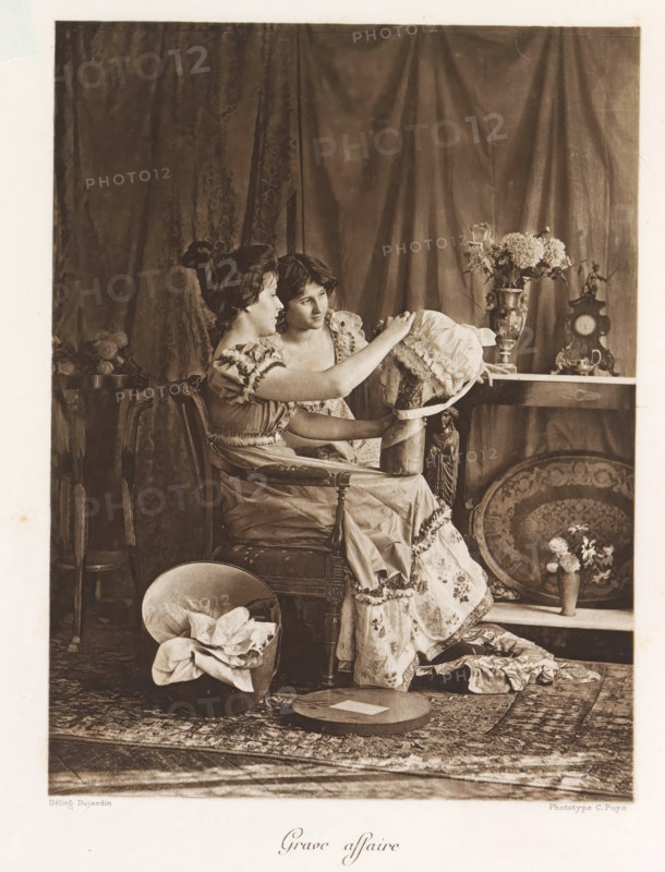 Grave affair, Two women look at a hat in an interior, page, photomechanical print, photographer: Constant Puyo, France, cliché maker: Paul Dujardin, printer (person): Charles Wittmann, Paris, c. 1886 - in or before 1896, printer (person), paper, photoaquatint, height 202 mm x width 150 mm, adult woman, fan, interior, representation of a building, Two young women sit closely on an upholstered chair in front of a draped curtain, one woman facing slightly sideways and the other leaning toward her while both focus on a bonnet that the nearer woman holds; the women wear patterned dresses with ruffled sleeves and layered skirts. Around them are decorative objects including a table with a tall ornate vase of flowers and an antique clock, a round decorative tray propped against the shelf, and a small pedestal with a figurine. On the floor in front of them a hat box contains folded fabric and a flat round hat box lid rests nearby on a patterned rug. The scene is sepia-toned with soft lighting a...