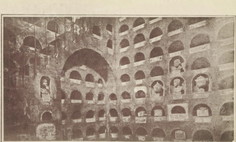 Interior of the columbarium of the Vigna Codini, Columbaria Tomb of 'The officers of Caesar's household', page, photomechanical print, number: 'Plate II', c. 1867 - in or before 1877, paper, photoaquatint, height 220 mm x width 140 mm, niche-tomb, wall-tomb, Rome, Columbarium of the Vigna Codini, A sepia-toned photograph of a wall filled with rows of recessed niches, many of which contain sculpted busts and urns, with several niches left empty; the wall shows arches and a large central archway, rough stone texture, and varying light and shadow across the surfaces, and a few vertical elements like poles or supports visible on the left side.