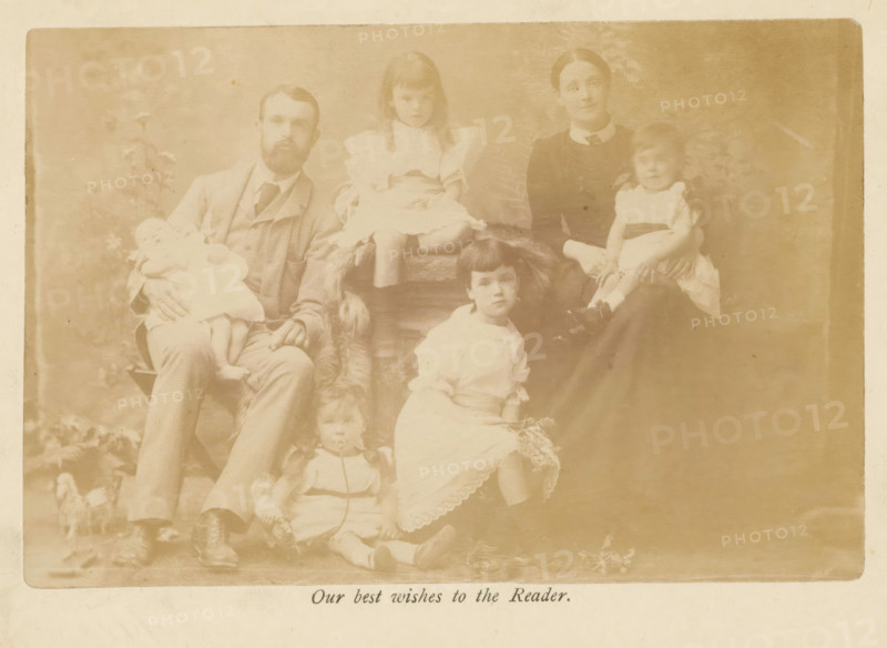 Group portrait of the family of Samuel Simmins, photograph, page, inscription: 'Our best wishes to the Reader.', c. 1882 - in or before 1887, photographer, paper, albumen print, albumendruk, height 95 mm x width 148 mm, Photo on frontispiece., Person, Samuel Simmins, family group, especially parents with their child(ren), anonymous historical persons depicted in a group, in a group-portrait, A faded sepia photograph showing a seated man with a beard wearing a suit and tie holding a small child in a light dress, a woman in a dark dress with a high collar seated nearby holding another small child in a light dress, and several children arranged around them wearing light-colored dresses and shoes; the background includes a painted backdrop with indistinct foliage and the image has a worn border.