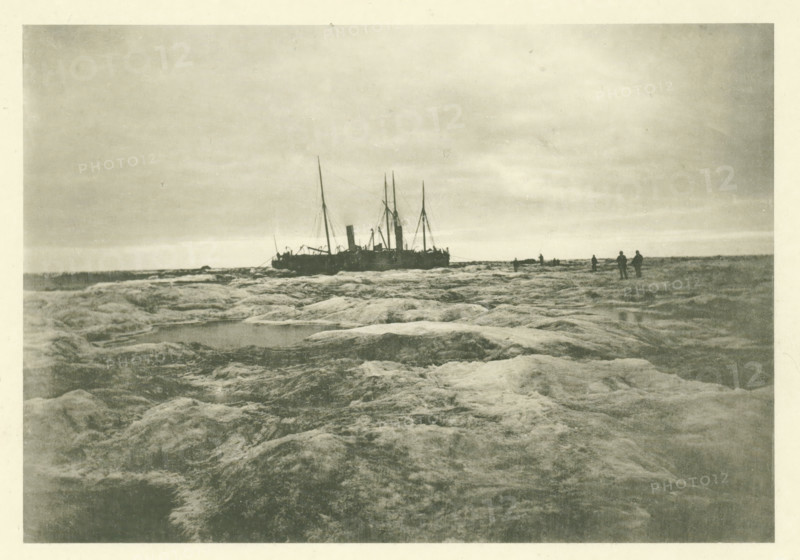 The Louise and the Varna moored in a patch of ice. Aug 17 1882, The Dutch Polar Expedition 1882-1883 published by the care of the Lieutenant Commander 1st class BJG Volck, photographer, photographer: H. Ekama, writer (clerical worker): Maurits Snellen, printer (person): E. Diener, London, publisher: Bosch & Zoon, LE, Balthasar Jean George Volck, c. 1886, writer (clerical worker), printer (person), paper, collotype, light printing on photo paper on cardboard, height 0 mm x width 0 mm, A grainy monochrome scene showing a shoreline with rough, textured rock surfaces in the foreground leading toward a distant horizon, a large ship with tall masts and a central smokestack positioned near the horizon line, several small human silhouettes standing on the rocks near the ship, a flat sky with subtle cloud banding above, and shallow pools of water among the rocky foreground.
