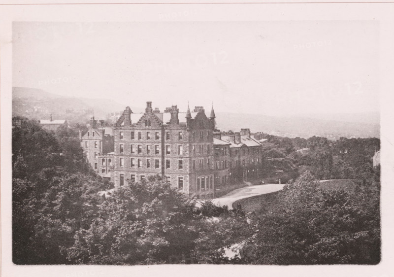 Craigslands hydropathic, page, photomechanical print, c. 1890 - c. 1900, paper, collotype, photoprint, height 101 mm x width 153 mm, medical examination, medical treatment, watering-place, spa, facade (of house or building), A sepia-toned photograph showing a large stone building with multiple stories, steeply pitched roofs, chimneys, and decorative gables, flanked by trees and shrubbery in the foreground and a curved driveway leading past the building, with additional smaller buildings extending to the right and a distant hazy landscape of hills and treetops in the background.