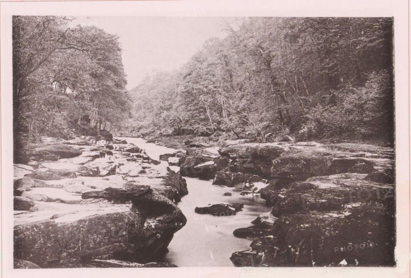 The 'Strid' on the ..., page, photomechanical print, c. 1890 - c. 1900, textile materials, linen (material), collotype, photoprint, height 102 mm x width 152 mm, river, A sepia-toned photograph of a rocky riverbed flowing away from the viewer, with smooth water weaving between large, flat rocks and boulders; dense trees and foliage line both banks, their branches arching over the scene, and the treeline recedes into a lighter, misty background.