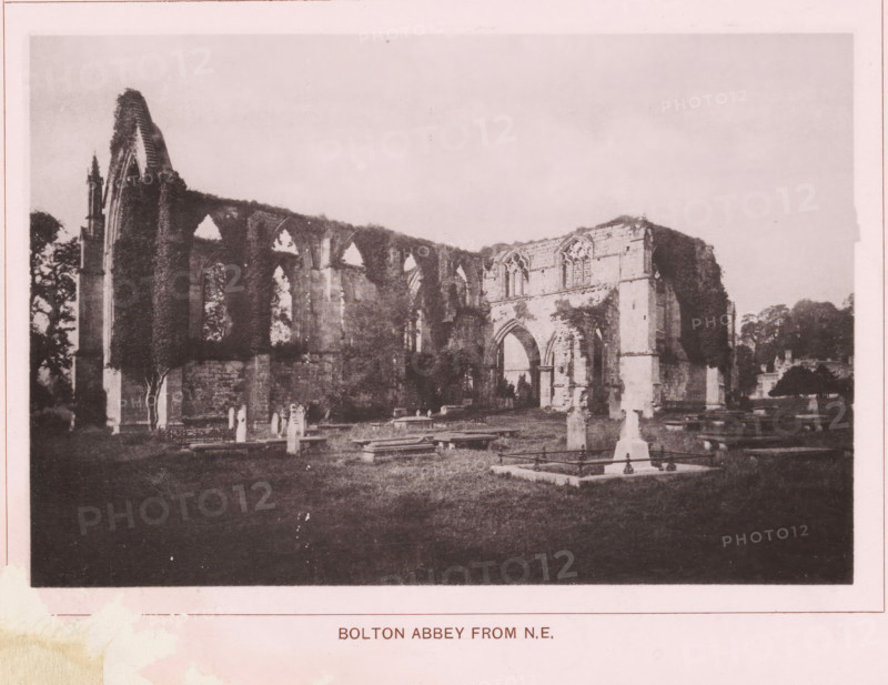 Bolton Abbey from NE, page, photomechanical print, c. 1890 - c. 1900, textile materials, linen (material), collotype, photoprint, height 102 mm x width 152 mm, ruin of church, monastery, Yorkshire, Wharfedale, Bolton Abbey, A sepia-toned photograph shows the ruins of a stone abbey with tall arched openings and partially collapsed walls surrounded by a grassy burial ground with upright and flat grave markers and low tombs; trees appear behind and beside the ruins and vegetation climbs parts of the remaining structure, while the sky above is pale and empty.