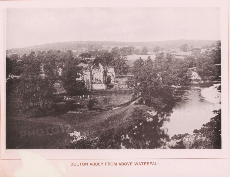 Bolton Abbey from above waterfall, page, photomechanical print, c. 1890 - c. 1900, textile materials, linen (material), collotype, photoprint, height 102 mm x width 152 mm, ruin of church, monastery, Yorkshire, Wharfedale, Bolton Abbey, A wide landscape photograph showing a river running along the right side with visible water flow and a lighter riverbank area, dense trees and foliage along both banks, open grassy land in the foreground, a large ruined stone building with a tall arched window and partial walls set among trees near the river, scattered small bright figures on the grass near the ruins, rolling wooded hills in the background, and a pale sky above.