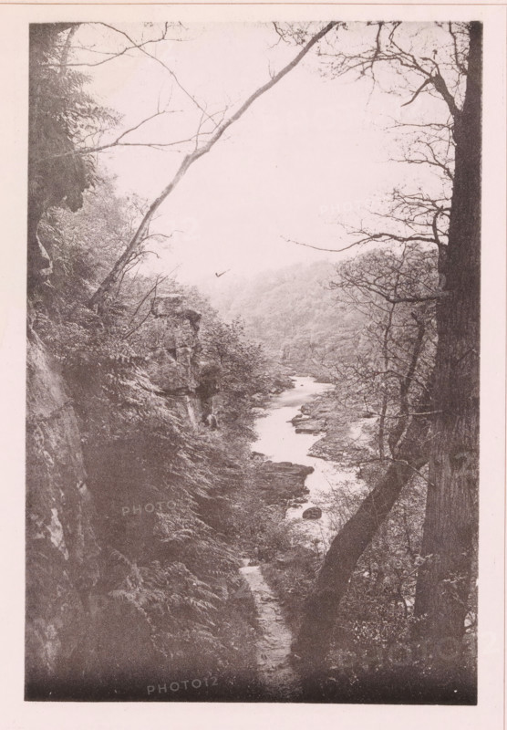 Bolton Hall, page, photomechanical print, c. 1890 - c. 1900, textile materials, linen (material), collotype, photoprint, height 102 mm x width 152 mm, country-house, façade (of house or building), North Yorkshire, A vertical grayscale photograph of a narrow river flowing through a rocky, wooded valley, framed by tree trunks and bare branches on both sides, with steep, vegetated slopes and exposed rock faces leading down to the water and distant misty hills visible in the background.