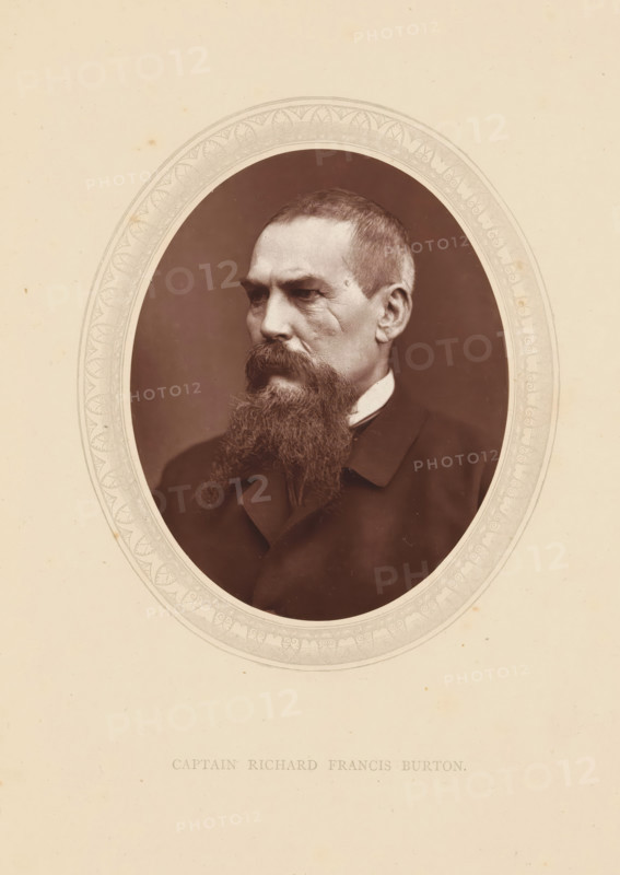 Captain Richard Francis Burton, page, photomechanical print, photographer: Lock & Whitfield, cliché maker: anonymous, c. 1871 - in or before 1876, paper, Woodburytype, height 115 mm x width 89 mm, Person, Richard Francis Burton, explorer, historical persons (portraits and scenes from the life) ((full) bust portrait), A sepia-toned oval portrait of a man with short hair and a long full beard, shown from the shoulders up and turned slightly to his left, wearing a dark coat and a high white collar, set against a dark background and surrounded by a pale ornate oval mat on a light paper.