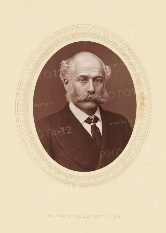 Sir Joseph William Bazalgette, page, photomechanical print, photographer: Lock & Whitfield, cliché maker: anonymous, c. 1871 - in or before 1876, paper, Woodburytype, Woodbury Process, height 115 mm x width 89 mm, Person, Joseph Bazalgette, engineering, historical persons (portraits and scenes from the life) ((full) bust portrait), A sepia-toned oval portrait set within a decorative oval border shows a middle-aged man with a receding hairline and bushy sideburns that connect to a thick mustache, wearing a dark suit jacket with a visible lapel, a white collared shirt, and a patterned tie; his expression is neutral and he faces forward against a plain dark background, with the surrounding paper showing subtle discoloration and a pale background.