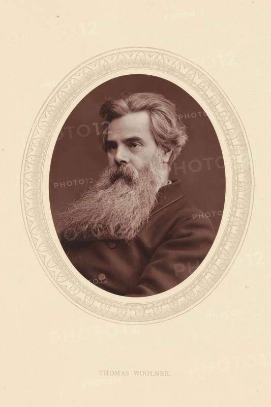 Thomas Woolner, page, photomechanical print, photographer: Lock & Whitfield, cliché maker: anonymous, c. 1871 - in or before 1876, paper, Woodburytype, Woodbury Process, height 115 mm x width 89 mm, Person, portrait, self-portrait of sculptor, historical persons (portraits and scenes from the life) ((full) bust portrait), A sepia portrait of a man shown from the chest up inside an ornate oval frame, with a full, long, bushy beard and thick wavy hair swept back, wearing a dark double-breasted coat, his face turned slightly to one side and his expression neutral, set against a plain dark background and mounted on a light-colored sheet with decorative border around the oval.