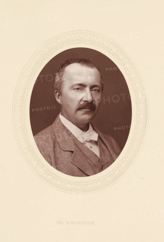 Dr. Schliemann, page, photomechanical print, photographer: Lock & Whitfield, cliché maker: anonymous, c. 1871 - in or before 1876, paper, Woodburytype, Woodbury Process, height 115 mm x width 89 mm, Person, Heinrich Schliemann, explorer, historical persons (portraits and scenes from the life) ((full) bust portrait), A sepia-toned oval portrait of a middle-aged man with short hair and a mustache, shown from the chest up and wearing a suit jacket, waistcoat, high-collared shirt, and a bow tie, set against a dark oval background and surrounded by a decorative light-colored oval frame on a pale rectangular sheet.