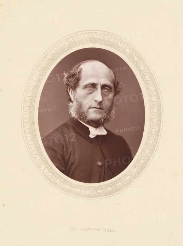 Rev. Newman Hall, page, photomechanical print, photographer: Lock & Whitfield, cliché maker: anonymous, c. 1871 - in or before 1876, paper, Woodburytype, Woodbury Process, height 115 mm x width 89 mm, Person, Christopher Newman Hall, historical persons (portraits and scenes from the life) ((full) bust portrait), A sepia-toned oval portrait of a middle-aged man with a high forehead and receding hairline, framed by sideburns that extend down into a full beard along his jaw, wearing a dark clerical coat with a visible button and a white clerical collar, set against a plain darker background and surrounded by a decorative oval mat with an intricate patterned border, all on a light cream paper with small spots and slight discoloration.