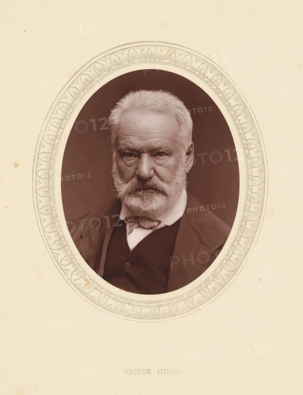 Victor Hugo, page, photomechanical print, photographer: Étienne Carjat, Paris, cliché maker: anonymous, c. 1871 - in or before 1876, paper, Woodburytype, Woodbury Process, height 115 mm x width 89 mm, Person, portrait of a writer, historical persons (portraits and scenes from the life) ((full) bust portrait), A sepia-toned oval portrait of an older man with light hair and a full beard and mustache, wearing a dark jacket and a light-colored shirt with a tied neck cloth, set against a dark background and surrounded by an ornate decorative oval border on a pale paper background.