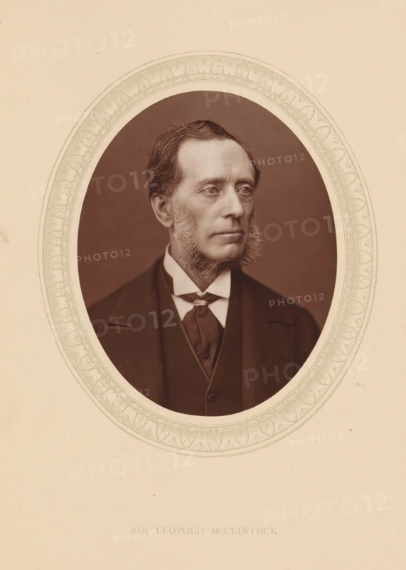 Sir Leopold McClintock, page, photomechanical print, photographer: Lock & Whitfield, cliché maker: anonymous, c. 1873 - in or before 1878, paper, Woodburytype, Woodbury Process, height 115 mm x width 89 mm, Person, Francis Leopold McClintock (sir), explorer, historical persons (portraits and scenes from the life) ((full) bust portrait), An oval-framed sepia portrait of a middle-aged man shown from the chest up, wearing a dark suit, waistcoat, high-collared shirt, and a cravat; he has short, neatly combed hair, prominent sideburns, and a composed facial expression while looking slightly to his right; the oval image is surrounded by a decorative patterned border set against a plain light background.