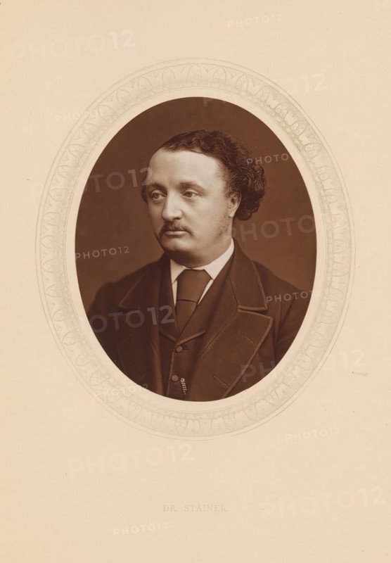 Dr. Stainer, page, photomechanical print, photographer: Lock & Whitfield, cliché maker: anonymous, c. 1873 - in or before 1878, paper, Woodburytype, Woodbury Process, height 115 mm x width 89 mm, Person, John Stainer, portrait of composer, historical persons (portraits and scenes from the life) ((full) bust portrait), A sepia portrait of a man shown from the chest up within an oval frame, wearing a dark suit jacket with wide lapels, a waistcoat, a white shirt, and a tie; he has short curly hair swept back, a mustache, and a contemplative facial expression, with a plain dark background inside the oval and a decorative patterned border around the oval set on a light beige background.