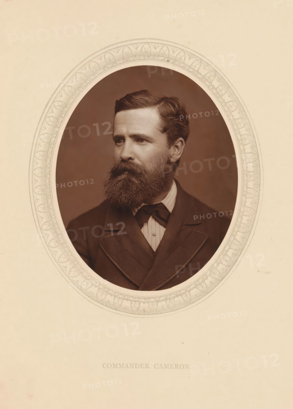 Commander Cameron, page, photomechanical print, photographer: Lock & Whitfield, cliché maker: anonymous, c. 1873 - in or before 1878, paper, Woodburytype, Woodbury Process, height 115 mm x width 89 mm, Person, Verney Lovett Cameron, explorer, historical persons (portraits and scenes from the life) ((full) bust portrait), A sepia-toned oval portrait of a man with a full beard and mustache, neatly combed hair, and a serious expression, wearing a dark suit jacket with wide lapels, a vest, a light-colored collared shirt, and a bow tie, set against a plain background and framed by an ornate patterned oval mat on a cream-colored page.