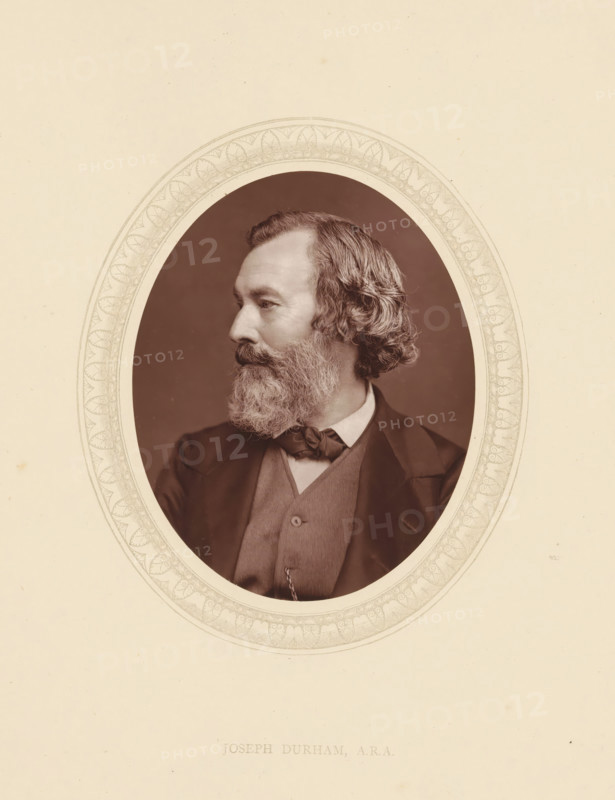 Joseph Durham, ARA, page, photomechanical print, photographer: Lock & Whitfield, cliché maker: anonymous, c. 1873 - in or before 1877-10-27, paper, Woodburytype, Woodbury Process, height 115 mm x width 89 mm, Person, Joseph Dunham, portrait, self-portrait of sculptor, historical persons (portraits and scenes from the life) ((full) bust portrait), A sepia oval portrait of a man shown in profile facing left, with wavy hair and a full beard, wearing a suit jacket, waistcoat, high-collared shirt, and bow tied at the neck, set against a plain dark background and framed by a decorative oval border on a light background.