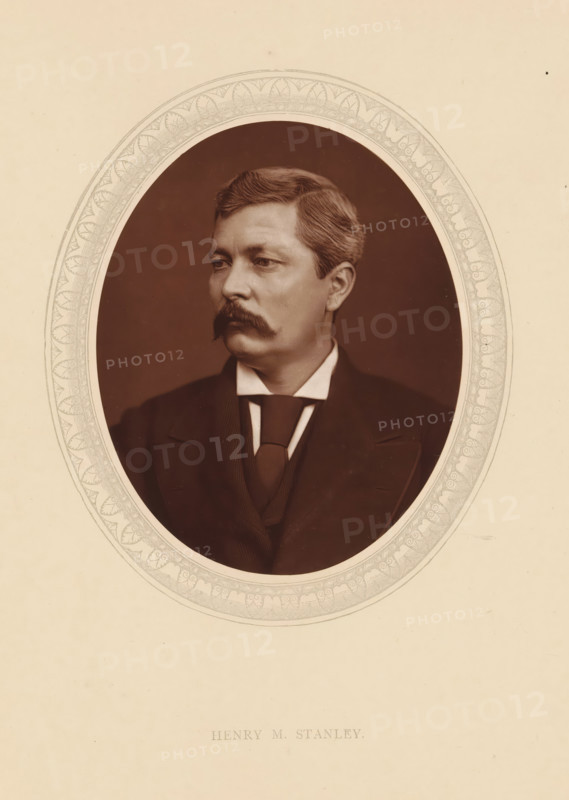 Henry M. Stanley, page, photomechanical print, photographer: Lock & Whitfield, cliché maker: anonymous, c. 1875 - in or before 1880, paper, Woodburytype, Woodbury Process, height 115 mm x width 89 mm, Person, sir Henry Morton Stanley, explorer, historical persons (portraits and scenes from the life) ((full) bust portrait), An oval sepia portrait of a man with neatly combed hair and a prominent mustache set against a dark background, framed by an ornate patterned border on a light beige sheet, the man wearing a dark suit jacket, waistcoat, high starched collar, and a tied cravat, his face turned slightly to the viewer’s left with a neutral expression.